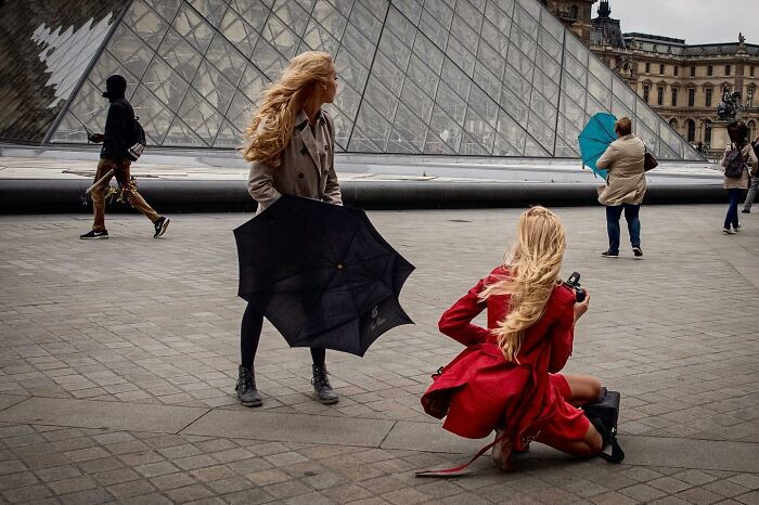 Street photography by Andrea Torrei: two women with windblown hair and umbrellas near a glass pyramid.