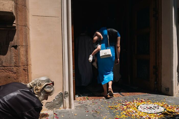 Street photo by Andrea Torrei showing a woman with a child entering a doorway, with petals on the ground nearby.