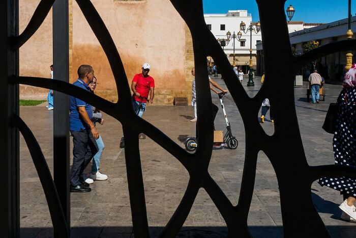 Street photo by Andrea Torrei showing people walking and a man with a scooter, viewed through a patterned metal structure.