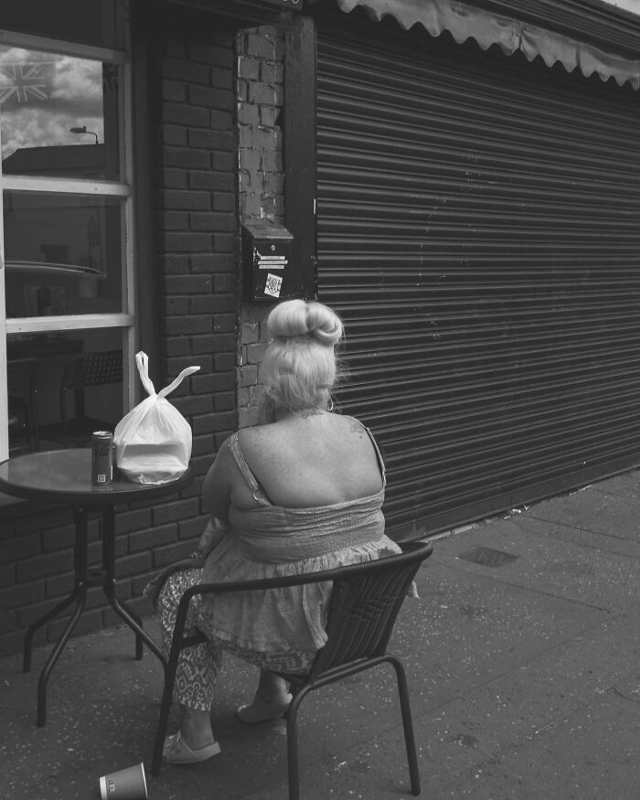 Street photo of a woman sitting by a shop with a bun hairstyle, viewed from behind.