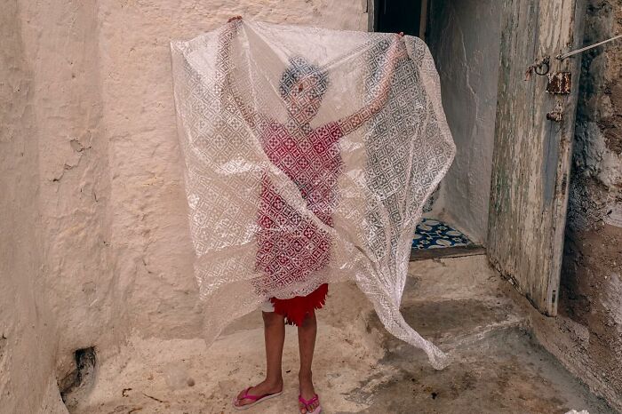 Child holding a delicate lace sheet in front of a rustic background, embodying curiosity in street photography.