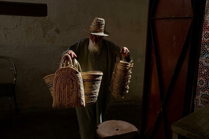 Elderly man with a woven hat holds handwoven baskets, standing in a dimly lit space.