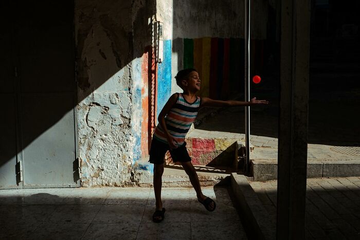 Child playing with a ball in the street, captured with a curious eye on world moments, colorful striped wall in the background.
