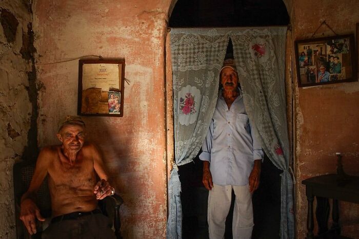 Two elderly men in an old room, one seated shirtless, the other standing, capturing street photo essence by Andrea Torrei.
