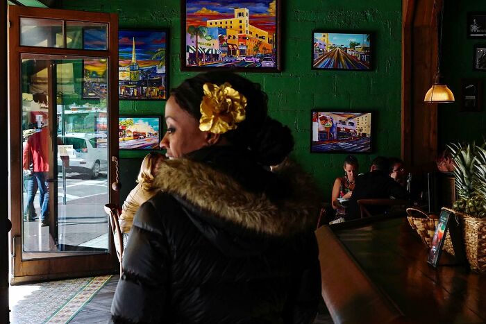 Street photo by Andrea Torrei: woman with yellow flower hair accessory in a cafe with vibrant paintings on the walls.