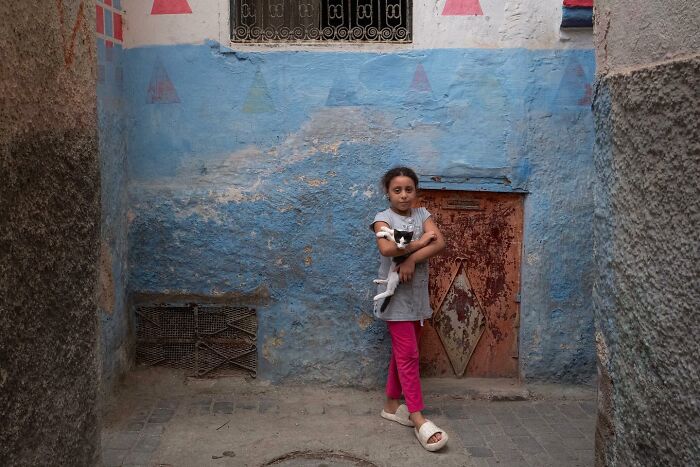 Girl in pink pants holding a doll, standing in a colorful alley, showcasing street photography by Andrea Torrei.