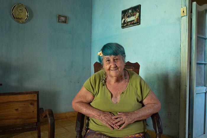 Elderly woman with blue hair in a green shirt sitting in a modest blue room. Street photo capturing a curious eye on the world.