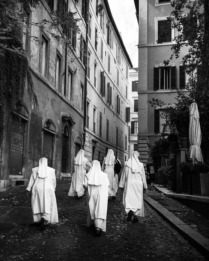 Nuns walking down a cobblestone street surrounded by historic buildings, captured in street photography by Andrea Torrei.