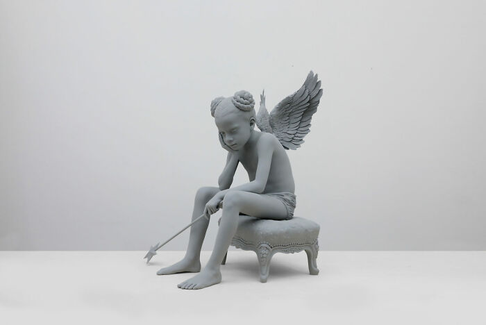 Monochrome sculpture by Hans Op De Beeck of a winged child sitting on a cushion, holding an arrow.