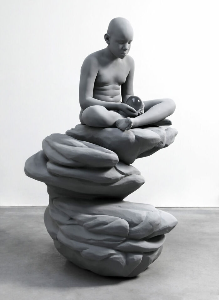 Monochrome sculpture by Hans Op De Beeck showing a seated figure on abstract rock formations, holding a reflective orb.