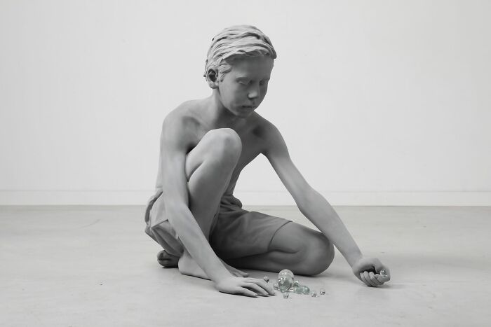Monochrome sculpture of a child kneeling, appearing contemplative, by Hans Op De Beeck.