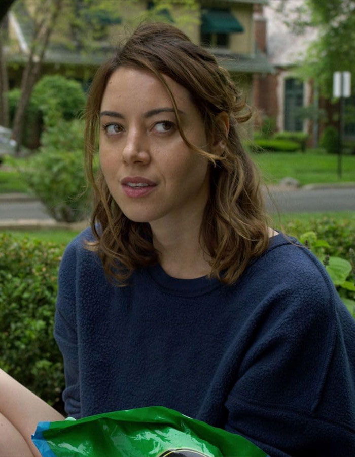 Woman sitting outdoors, wearing a blue sweater, with trees and houses in the background.