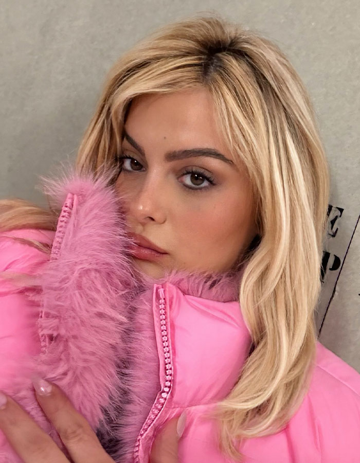 A woman in a pink fur-trimmed jacket looks into the camera.
