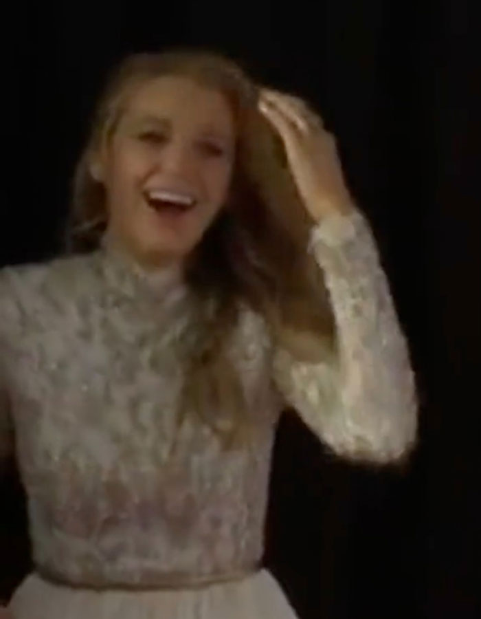 Blake Lively in a detailed lace dress, smiling and touching her hair against a dark background on the red carpet.