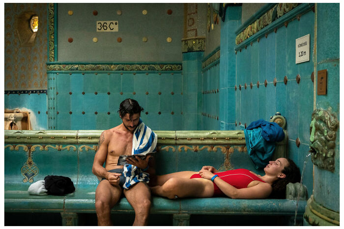Man reading on a tablet while a woman in a red swimsuit relaxes in an ornately tiled bath space, captured by Andrés Ramos.