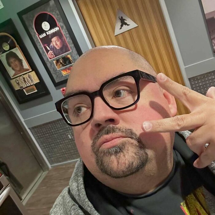 Celebrity posing indoors with peace sign, wearing glasses, framed posters behind.