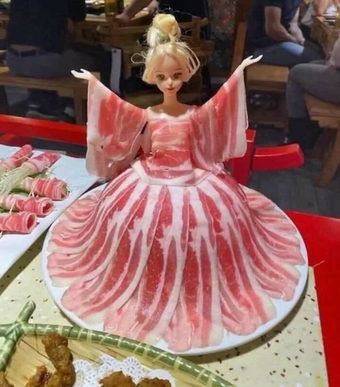 A doll dressed in a gown made of bacon slices, showcasing chaotic food creativity on a plate.