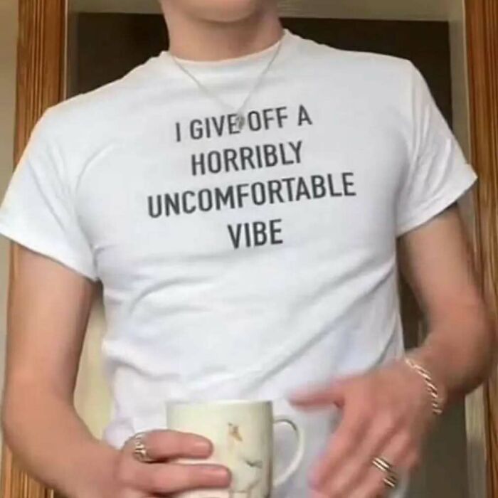 Person in white T-shirt with text saying "I give off a horribly uncomfortable vibe," holding a mug. Chaotic T-shirts theme.