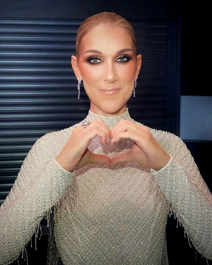 Celebrity in a sparkling outfit making a heart shape with hands, highlighting extravagant style and charm.