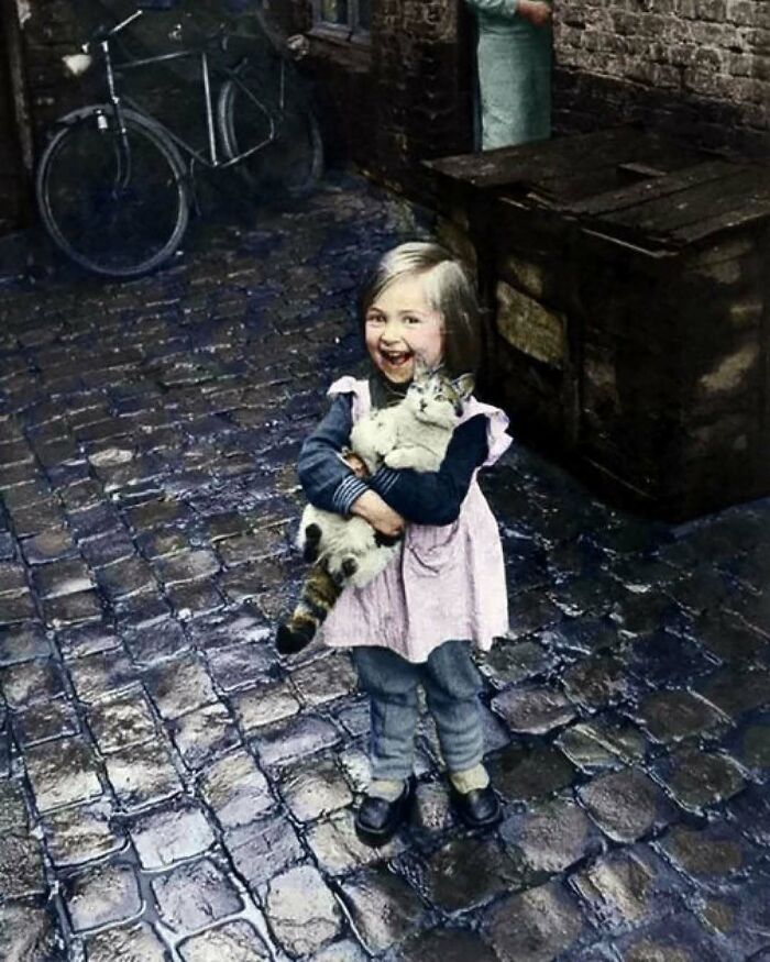 Young girl in historical attire stands on a cobblestone street, smiling and holding a cat, with a bicycle in the background.