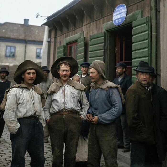 Men standing outside a wooden building wearing hats and old work clothes, capturing a historical moment from the past.