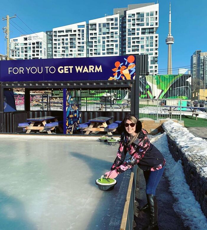 Woman in ear muffs playing outdoor curling in Canada with city skyline in the background.
