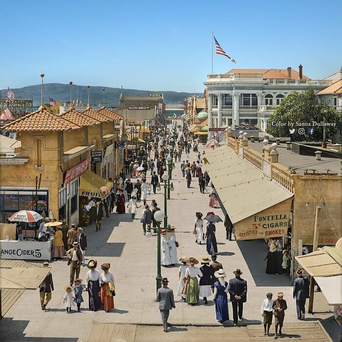 Bustling historical street scene with people in early 1900s attire, vibrant storefronts, and an American flag in the background.