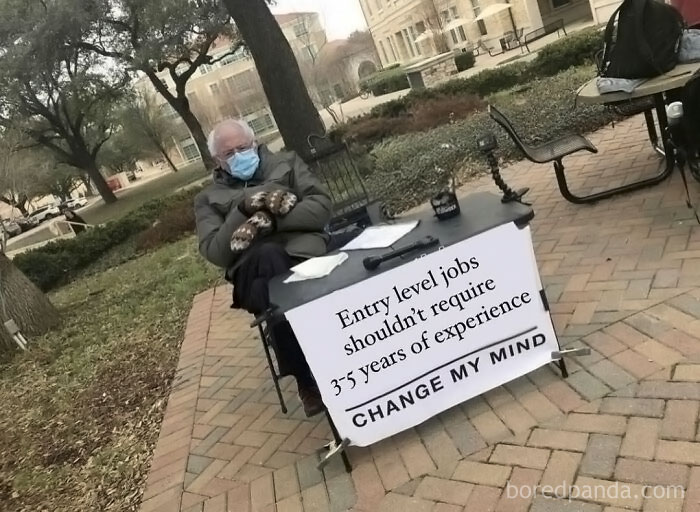 Person at a table with a sign on work-related humor about entry-level job experience, in an outdoor setting.