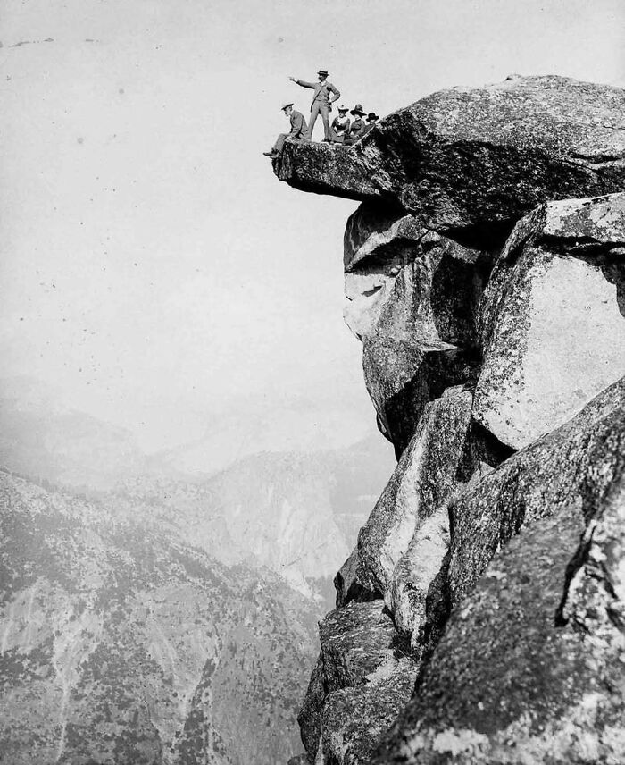 Historical picture of four men standing on an overhanging rock, overlooking a vast mountainous landscape.