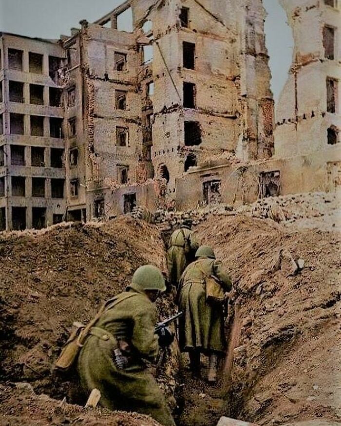 Soldiers in WWII uniforms advance through trenches amidst ruined buildings, highlighting historical wartime scenes.