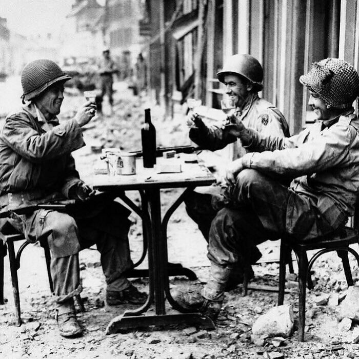 Three soldiers in historical attire share a drink, sitting outdoors at a small table in a wartime setting.