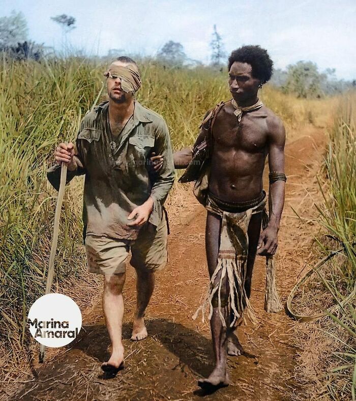 Blindfolded soldier assisted by local wearing traditional attire on a dirt path, showcasing historical moments of the past.