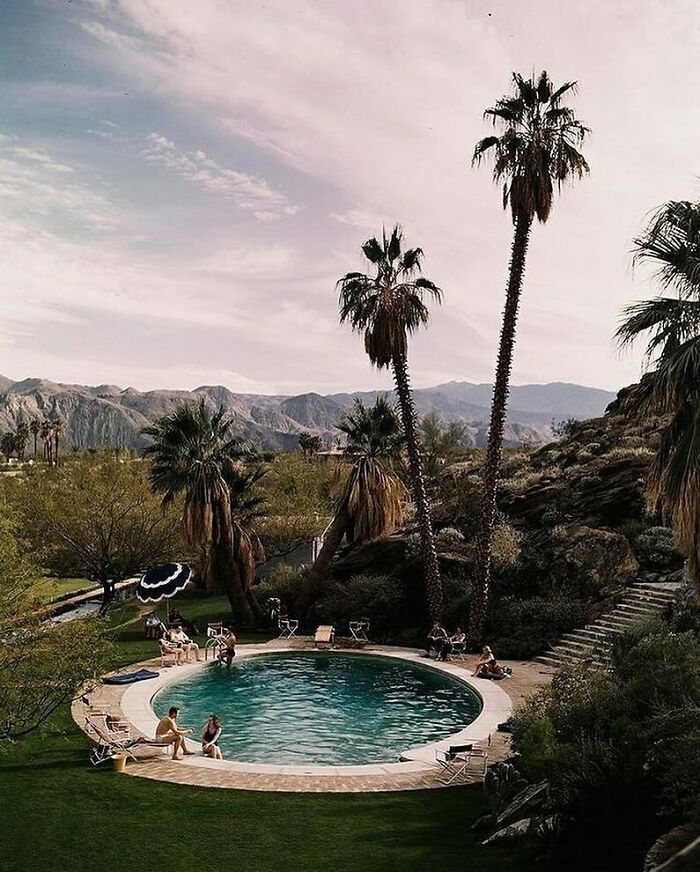 Vintage scene of people relaxing by a round pool with palm trees and mountains in the background.