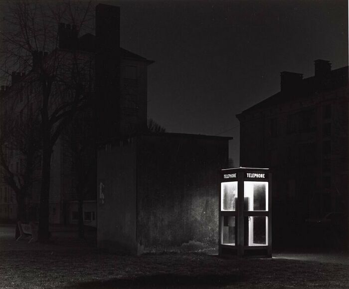 Historical photo of a glowing telephone booth at night, surrounded by dark buildings.