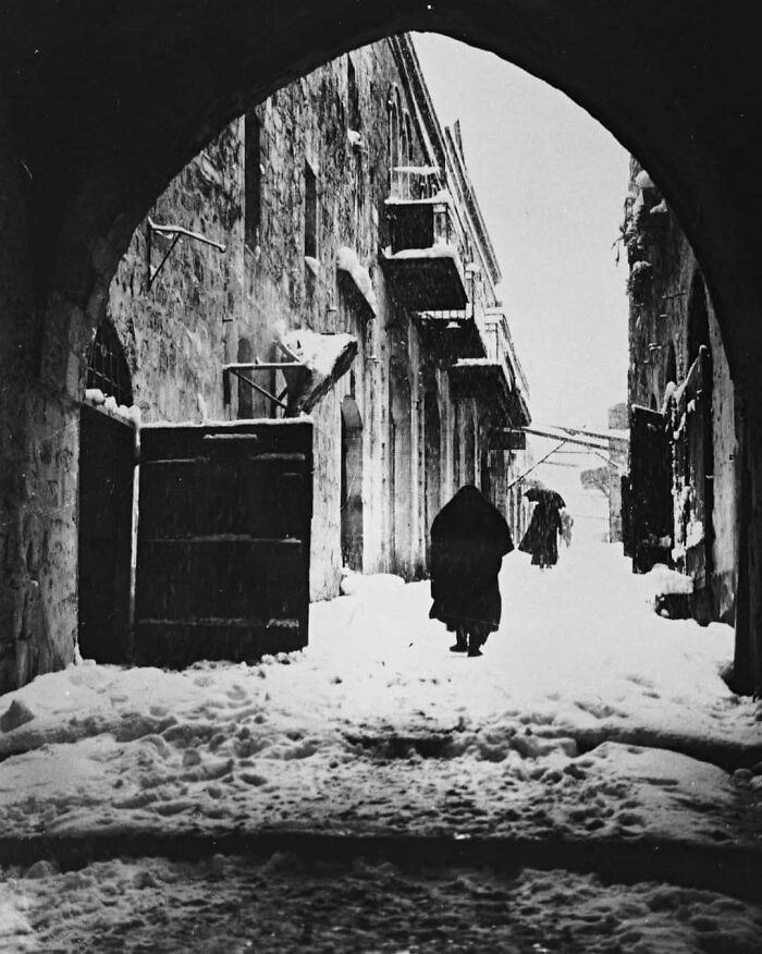 Snow-covered historical street scene under an archway, featuring a lone figure walking, evoking nostalgia.