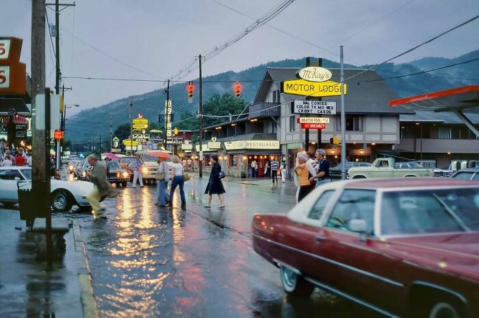 Vintage street scene with people crossing, classic cars, and a nostalgic 1960s motel sign in a rainy evening setting.