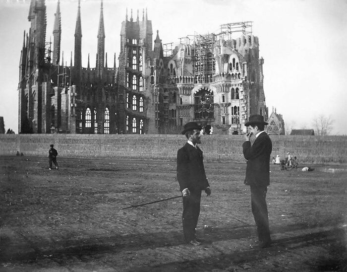 Two men in suits and hats converse in front of a partially constructed historical building, evoking nostalgia.