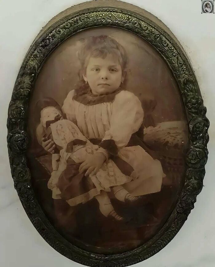 Antique headstone portrait of a young girl holding a doll, set in an ornate oval frame.