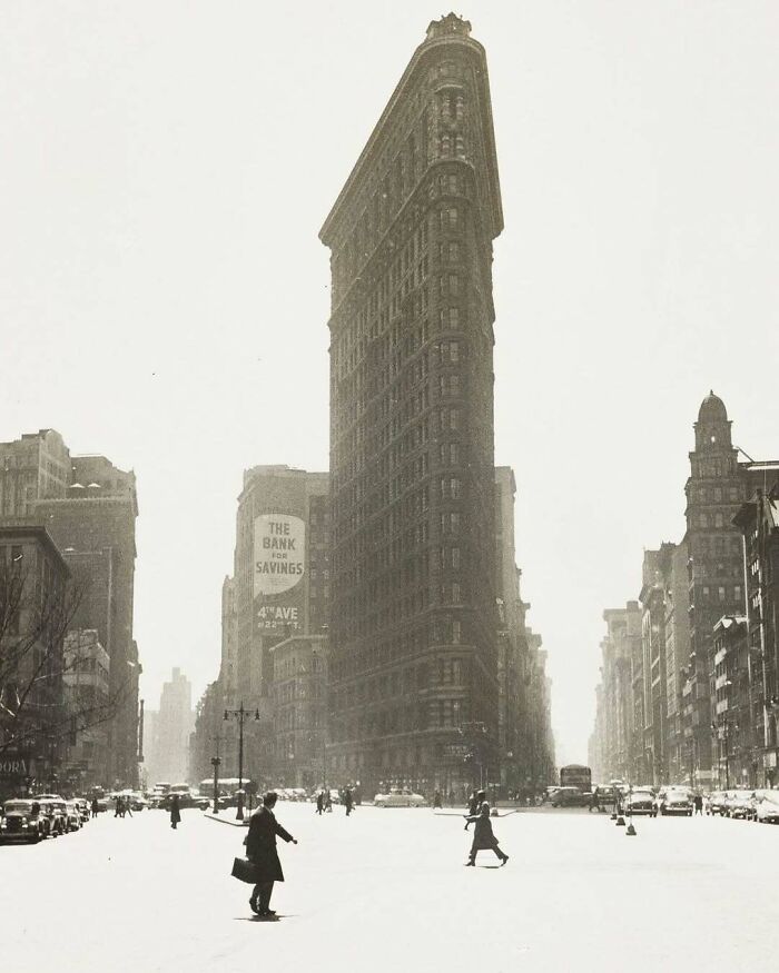 Historical pic of the Flatiron Building in a nostalgic cityscape scene with pedestrians and vintage cars.