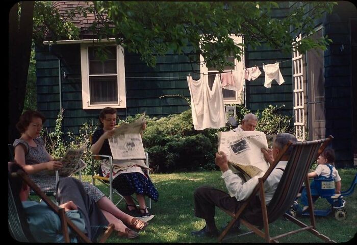People relaxing in a garden, reading newspapers, with historical nostalgia. A child sits nearby, and laundry hangs in the background.