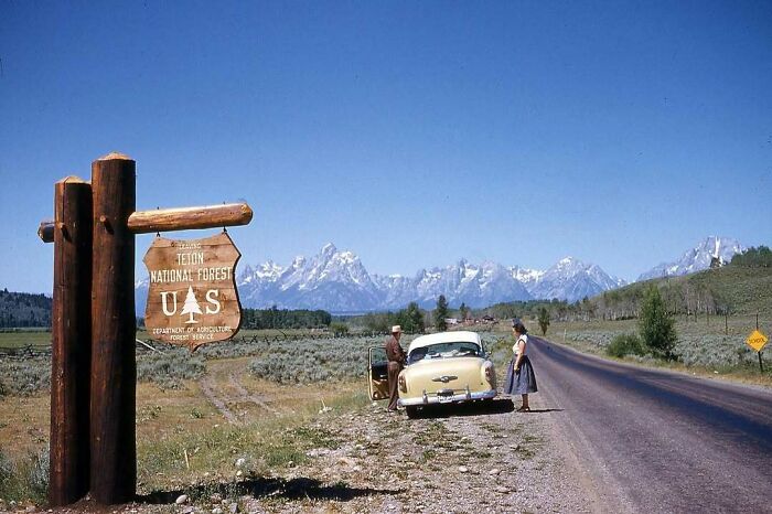Vintage photo of a couple by a car near Teton National Forest sign, with scenic mountain view. Historical nostalgia.