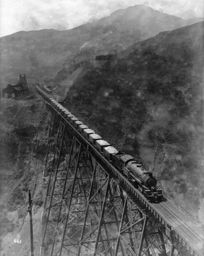 Historic steam train crossing a tall bridge, set against mountainous terrain.