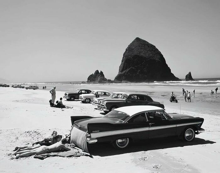 Vintage cars parked on a sandy beach with people sunbathing, evoking nostalgia of past seaside outings.