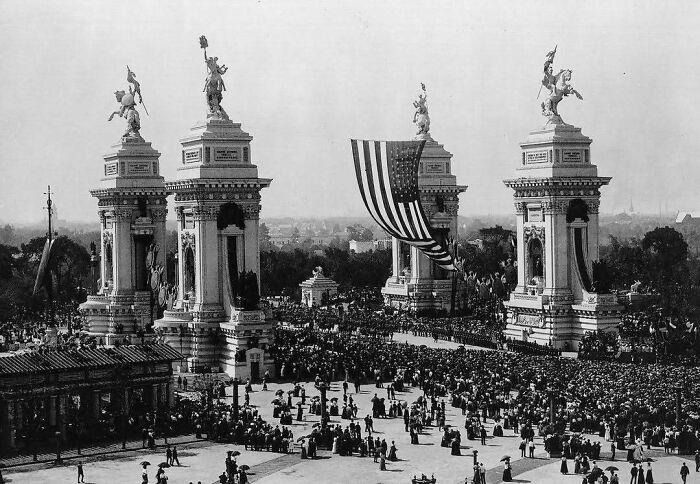 Crowds gather around historical architecture with statues and a large American flag draped between pillars.