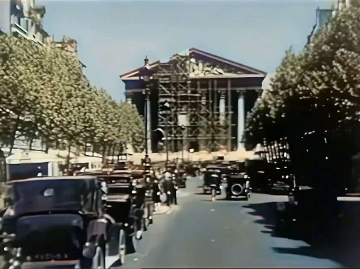 Historical street scene with vintage cars lined up under trees, leading to a classical building.
