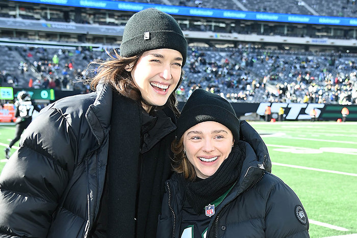"Chloë Grace Moretz and Kate Harrison smiling at a football game, both wearing black beanies and jackets."