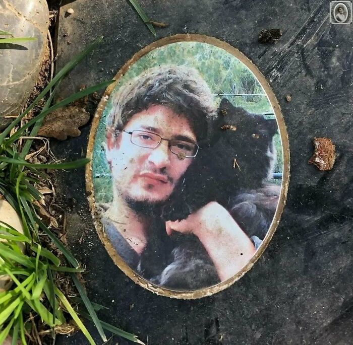 A headstone portrait of a person with glasses holding a black cat, surrounded by grass and stones.