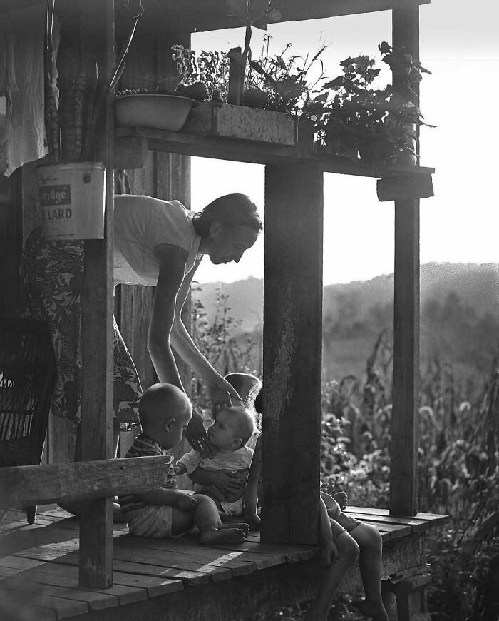 A nostalgic historical scene of a woman tending to children on a rustic porch, evoking a sense of timeless simplicity.