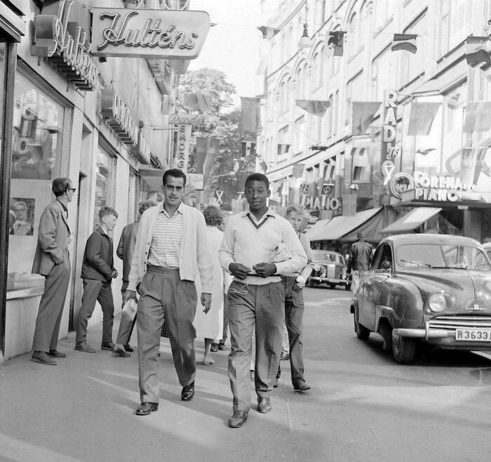 Historical street scene with people walking, vintage cars, and old shop signs, capturing urban nostalgia.
