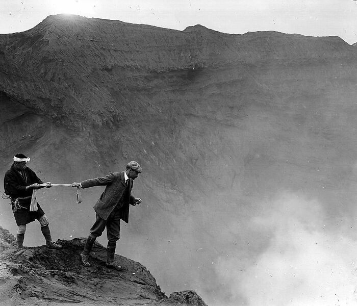Two men at the edge of a crater, one peering inside, creating a nostalgic historical scene.